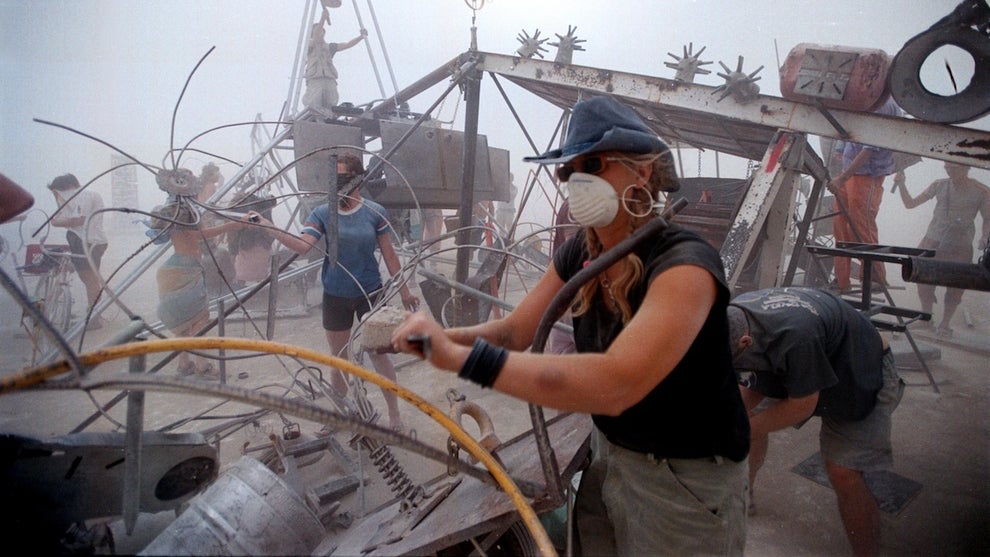 A complex desert vehicle is serviced as people working around it wear face masks.