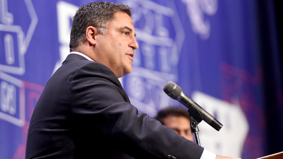 A man with medium-brown skin stands in front of a microphone at a podium. There is a blue background with blurry text on it, and part of the head of a person in the background.