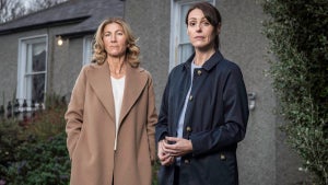 Two women in jackets stand in front of a modest home, with ivy on either side. They are both women with light-toned skin. The one on the left wears a tan coat, while the one on the right wears a navy jacket.