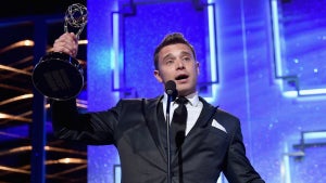 A man with light-toned skin holds up a gold trophy of a woman holding a globe-like structure, in front of a purple background. The man wears a suit and speaks in front of a microphone.