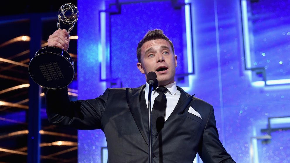 A man with light-toned skin holds up a gold trophy of a woman holding a globe-like structure, in front of a purple background. The man wears a suit and speaks in front of a microphone.