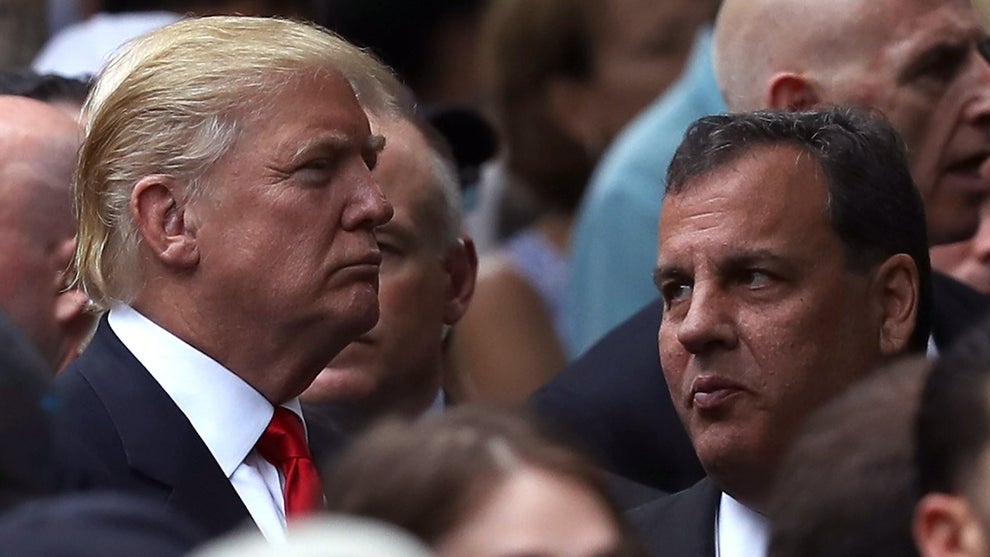 A man stands looking somber on the left, he has light-toned skin and light blonde hair. A man on the right looks toward the man on the left, while most of the crowd looks in the same direction as the man on the left. The man on the left is Donald Trump while the one on the right is Chris Christie.