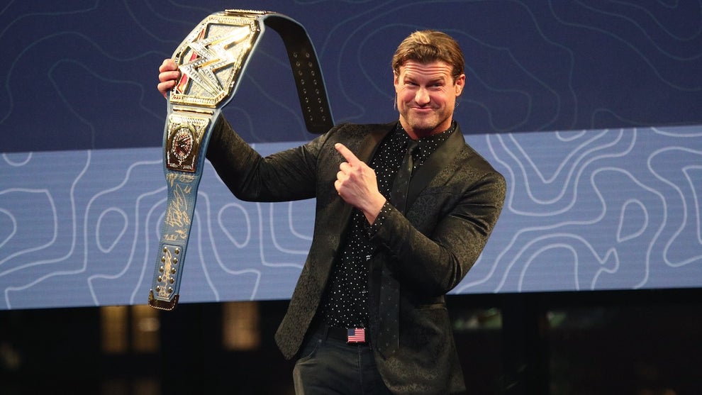 A pro wrestler wears a blazer and holds up a gold title belt. He has blonde hair.