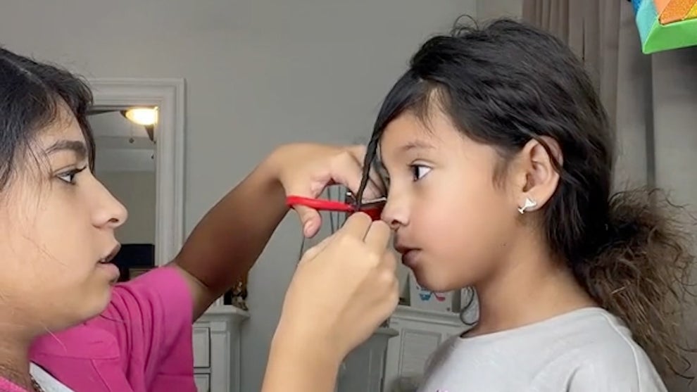 A girl in a pink jacket holds a pair of sisters, cutting her sister's hair. They both have light brown skin.