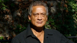 A man with deep brown skin wears an open-collar black shirt, in front of ivy on a building. He has dark gray hair.