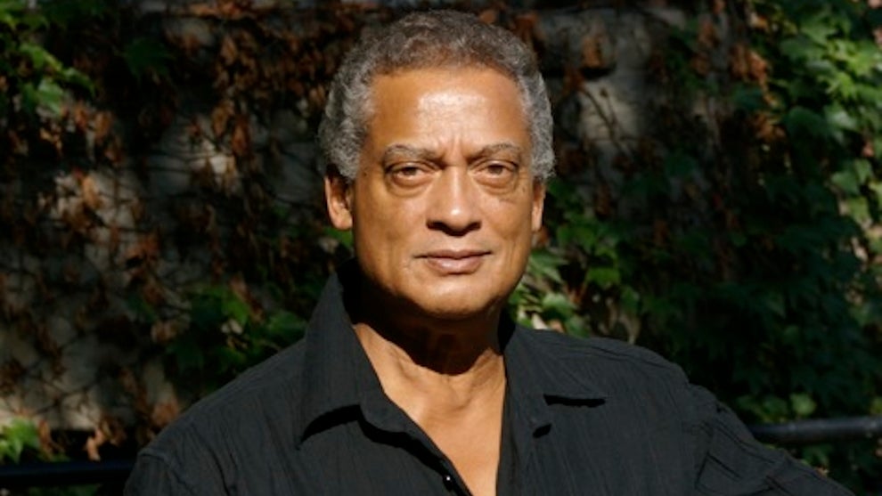 A man with deep brown skin wears an open-collar black shirt, in front of ivy on a building. He has dark gray hair.