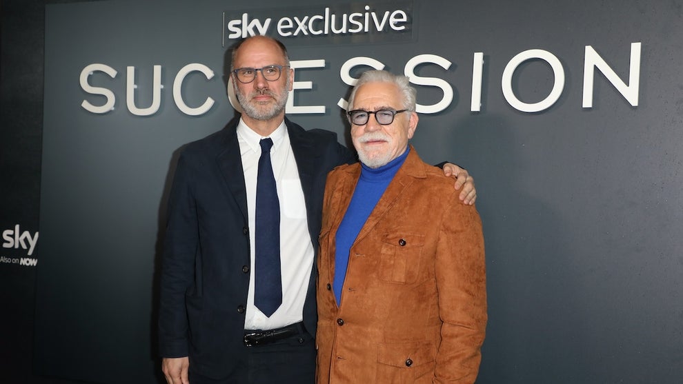 A middle-aged white man puts his arm around an older white man. They both wear suits, though the man on the right wears a bolder deep tan blazer. They stand in front of a background reading "Sky Exclusive: Succession."