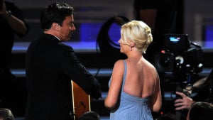 Jimmy Fallon and Amy Poehler viewed from behind, two adults with light skin. Fallon wears a suit and plays an acoustic guitar, while Poehler wears a dress with an open back. They look at each other and are standing in front of a seated crowd that's in the foreground.