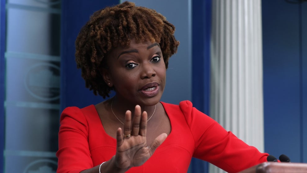A Black woman holds up her hand while making an animated, skeptical facial expression. She wears a red top and stands on a White House stage with a white column and blue background behind her.