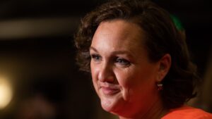 Rep. Katie Porter, a light-skinned woman with an orange top and wavy brown hair, looks off-camera.