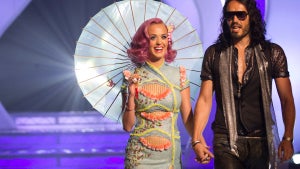 Katy Perry in a seafoam-green dress and carrying a parasol holds hands with Russell Brand, who wears a deep-V top, sunglasses, and a scarf as they walk out onto a stage.