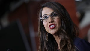 A woman with light-toned skin speaks emphatically. She wears glasses and bright red lipstick and has long brown hair. She speaks in a formal setting with apparent wood paneling in the background.
