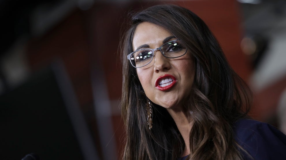 A woman with light-toned skin speaks emphatically. She wears glasses and bright red lipstick and has long brown hair. She speaks in a formal setting with apparent wood paneling in the background.