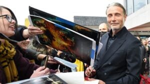 Mads Mikkelsen, a light-skinned man with silver hair and a scruffy beard, looks at the camera as he signs autographed posters for a group of nearby fans