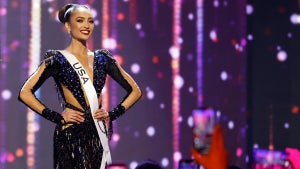 Miss USA contestant with medium-brown skin, wearing a dress with cutouts on the arms, stomach and chest, wearing a USA sash and smiling toward the camera. She stands in front of a purple sparkly backdrop. People in the foreground hold up cameraphones