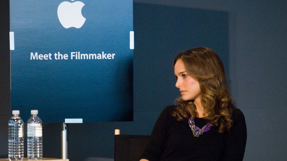 Natalie Portman sits in a chair looking to her left, where a sign in the background reads "Meet the filmmaker" beneath an Apple logo, shaped like an Apple fruit with a bite out of it. She has light brunette hair and wears a black top. There are two water bottles on a nearby table. She holds a microphone in her lap.
