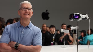 A man in a sky blue polo shirt stands in front of a small gathering of people. An Apple logo is on the wall in the back. A pair of augment reality goggles is on a stand to the right.