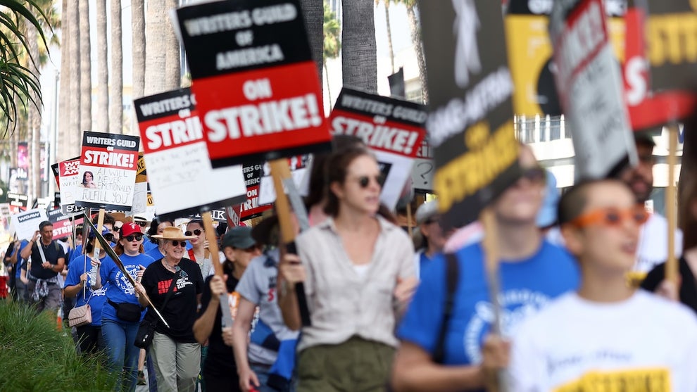 A large number of people walk down a sidewalk, holding picket signs saying "Writers Guild of America ON STRIKE!"