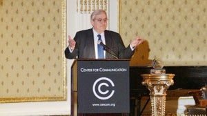 A man in a suit stands at a lectern and gestures as he speaks.