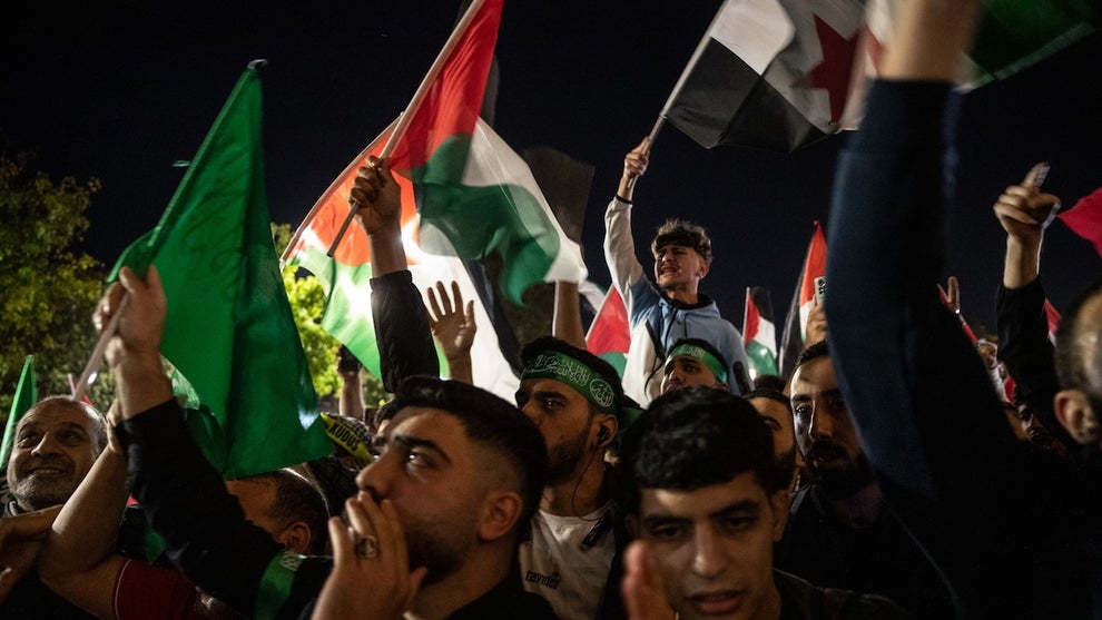 A group of men with medium-toned skin hold up flags and shout at a crowded gathering