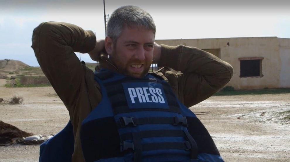 A man with gray hair adjusts the straps behind his head of a bullet proof vest that says "Press" on it.