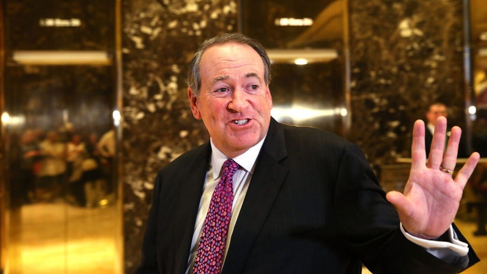 A man in a black suit with a red tie gestures.