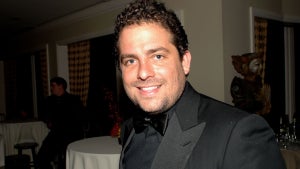 A man with curly black hair smiles at the camera. He has light-toned skin. He wears a black suit with black shirt and bowtie in a room with round tables with tablecloths.