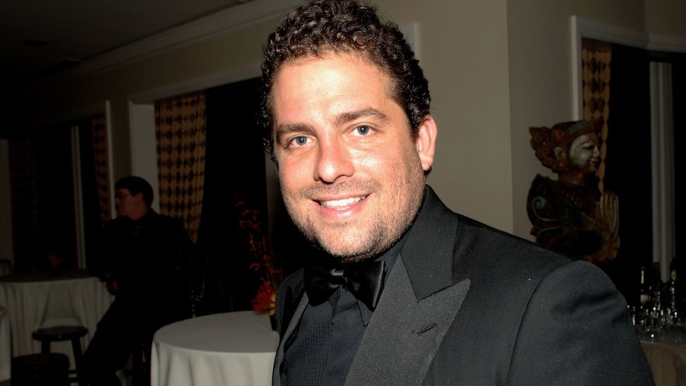 A man with curly black hair smiles at the camera. He has light-toned skin. He wears a black suit with black shirt and bowtie in a room with round tables with tablecloths.