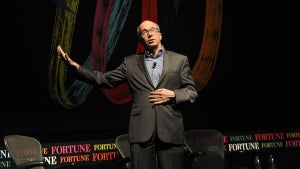 Fortune CEO Alan Murray speaks. He's an older man with light-toned skin in a gray suit and he's gesturing on a stage with a strip of the text "Fortune" behind him and several large colorful ovals.
