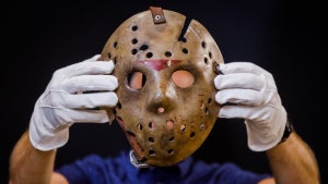 A person wearing white gloves holds up a weathered Jason Voorhees hockey mask.
