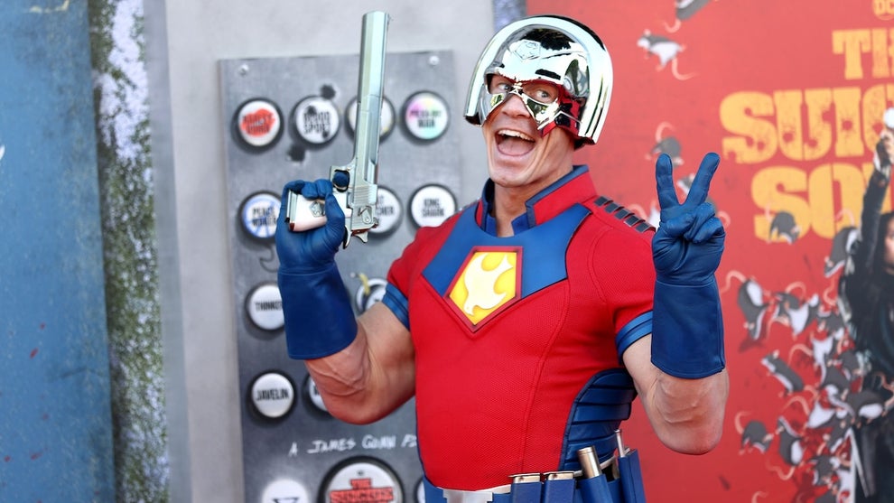 Pro wrestler and actor John Cena in costume as Peacemaker, with a red top, blue gloves, holding a long gun, wearing a silver helmet, smiling and making a peace sign. He appears on a red carpet with a backdrop for "The Suicide Squad."