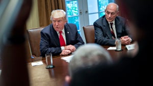 Two men with light-toned skin set at a large conference table, across from people whose faces are unseen in the foreground. The man on the right looks to the man on the left, who wears a bright red tie and appears to be speaking. The man on the left is then-President Donald Trump.