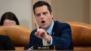 A white man with an upset expression holds up his finger. A nameplate in front of him reads. "MR. GAETZ." He wears a suit and is seated, with a couple others to his right.