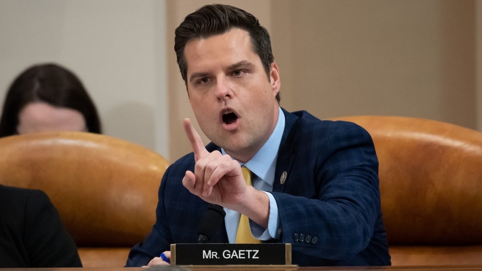 A white man with an upset expression holds up his finger. A nameplate in front of him reads. "MR. GAETZ." He wears a suit and is seated, with a couple others to his right.