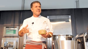 A chef holds a container with food in it, wears a chef jacket, and stands in front of cookware in a kitchen. He has light-toned skin.