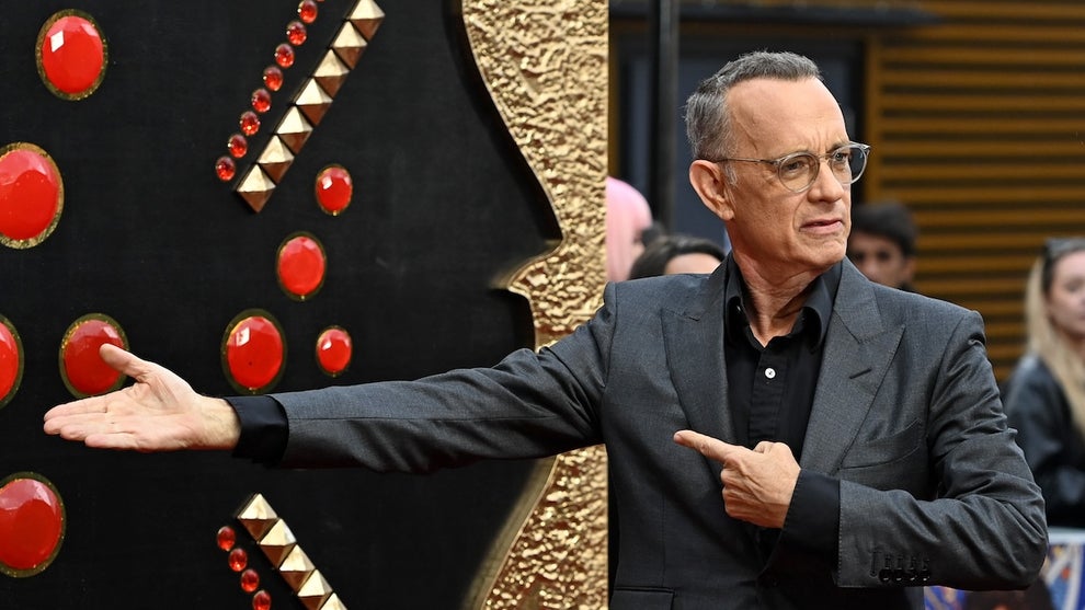 A man in a suit points toward a giant backdrop that appears to have elements resembling giant bedazzled jewels, with a serious expression on his face. He has light-toned skin and there are other people in the background.