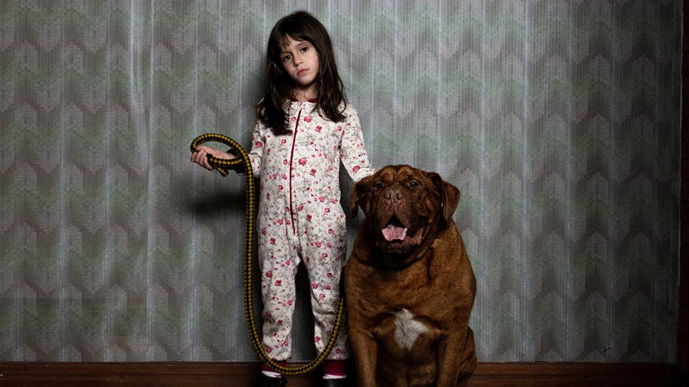 A little girl stands next to a large dog, holding his leash and looking direct into camera in still photo from "When Evil Lurks"