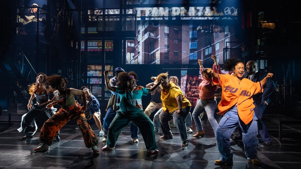 A group of people, mostly with dark-toned skin, dance on stage with a faux city background.