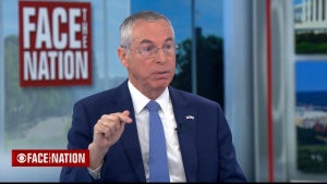 A light-skinned man in a suit points, gesturing, on a TV set. A sign behind him reads "FACE THE NATION."