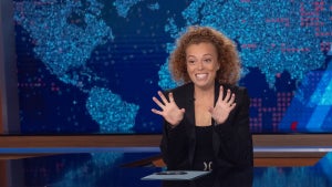A woman with medium-light-toned skin holds up both hands, as if grabbing breasts, while sitting at a desk in front of a backdrop showing a slightly abstract depiction of a map of the Earth.