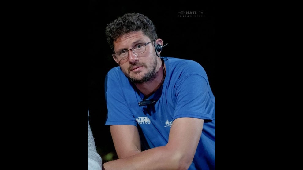 A man with medium-light-toned skin leans forward, wearing a blue T-shirt and glasses, as well as an earpiece.