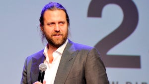 A light-skinned man with long stringy hair and a beard, wearing an open-collar shirt and a gray blazer, holds a microphone.