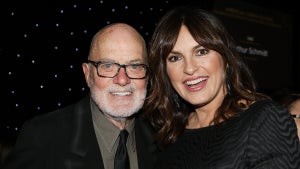 An older man and a woman stand next to each other, smiling and showing their teeth. He wears a suit, she wears a black top with gray waves on it. They are in front of a background of a faux night sky.