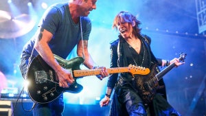 A light-skinned man and a medium-toned-skinned man play guitars on stage. The man on the right makes an extreme facial expression.
