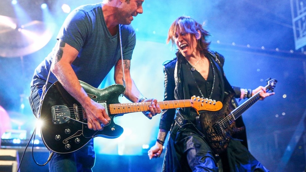 A light-skinned man and a medium-toned-skinned man play guitars on stage. The man on the right makes an extreme facial expression.