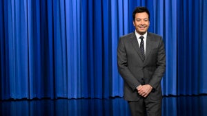 A man with light-toned skin in a suit in front of a blue curtain on a shiny black stage.