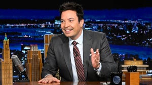 A man with an enthusiastic expression gestures while sitting at a desk, looking to the side. He has medium-light-toned skin and wears a suit.