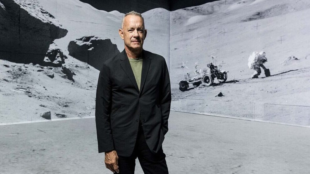 Actor Tom Hanks stands in front of a large projection of astronauts walking on the moon. The Comebacker