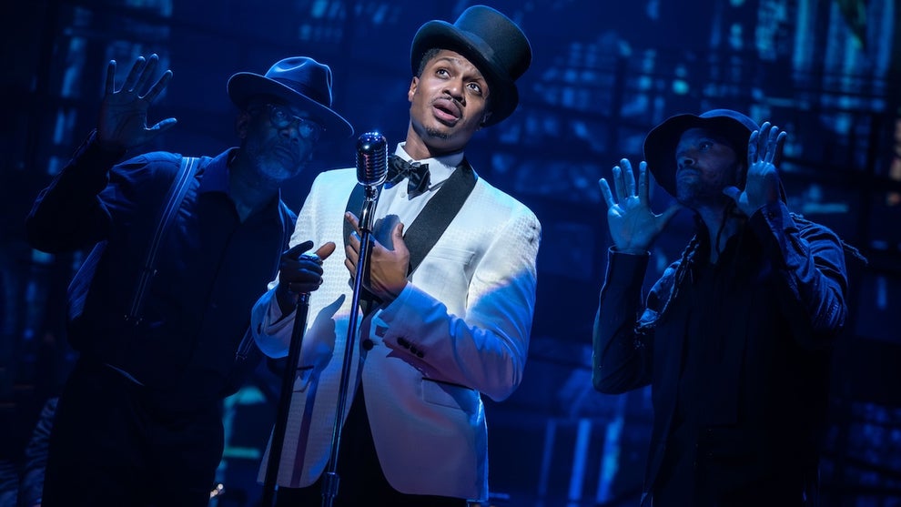 A Black man in a white suit jacket with bowtie and a top hat stands on stage in front of a microphone, two others holding up their hands in the shadows behind him.