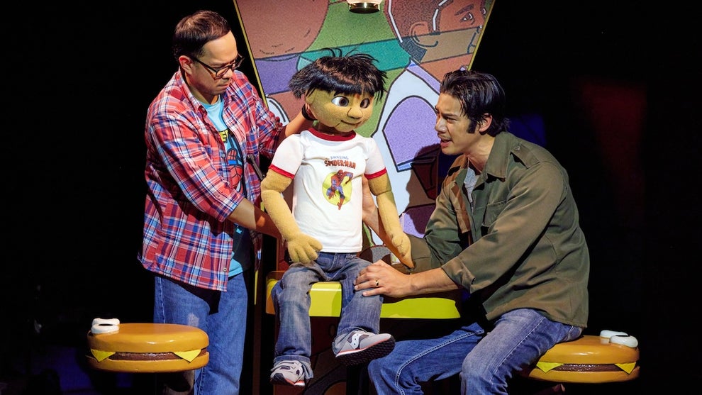 Two Asian men flank a puppet with similarly-toned skin, which appears to be a child wearing a Spider-Man T-shirt. They sit on hamburger bun-shaped stools.